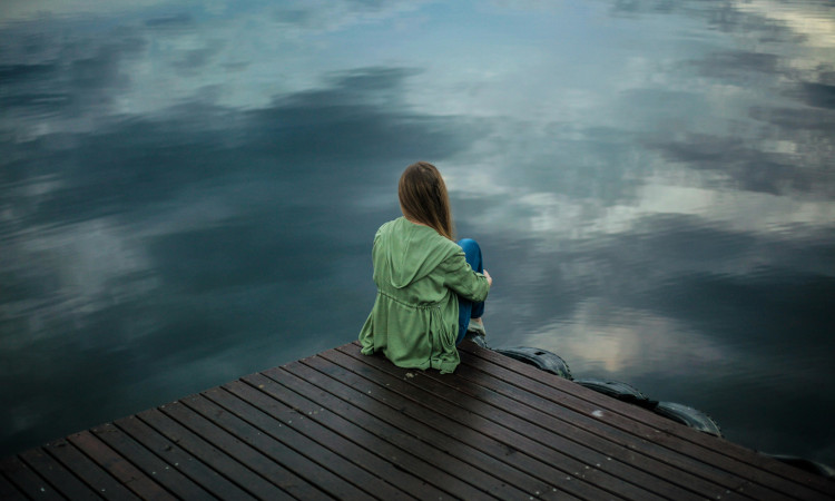 Mulher sentada num estrado de madeira a olhar para um lago, com um casaco verde vestido e a pensar nos sintomas de depress&atilde;o.