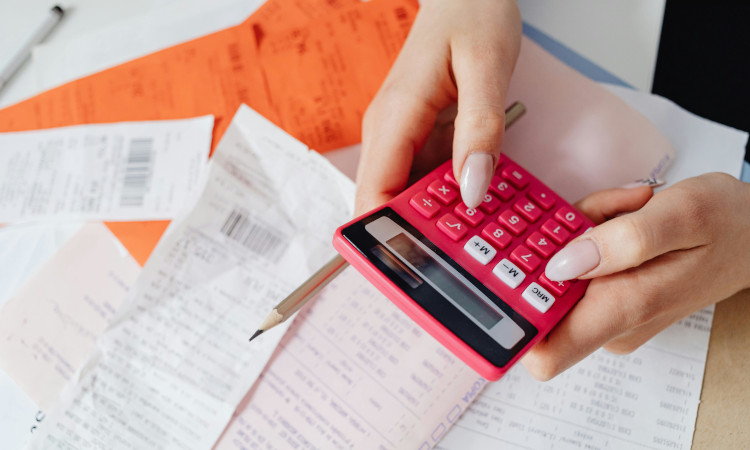 Mãos de mulher de unhas grandes em tons rosados a mexer em calculadora e com faturas em baixo a pensar como emitir Recibos Verdes.