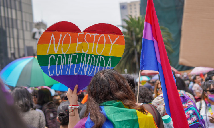Pessoas na rua com uma mulher em destaque, a segurar um cartaz colorido em forma de coração que diz "Não estou confusa" em espanhol, para ilustrar o que é ser bissexual.