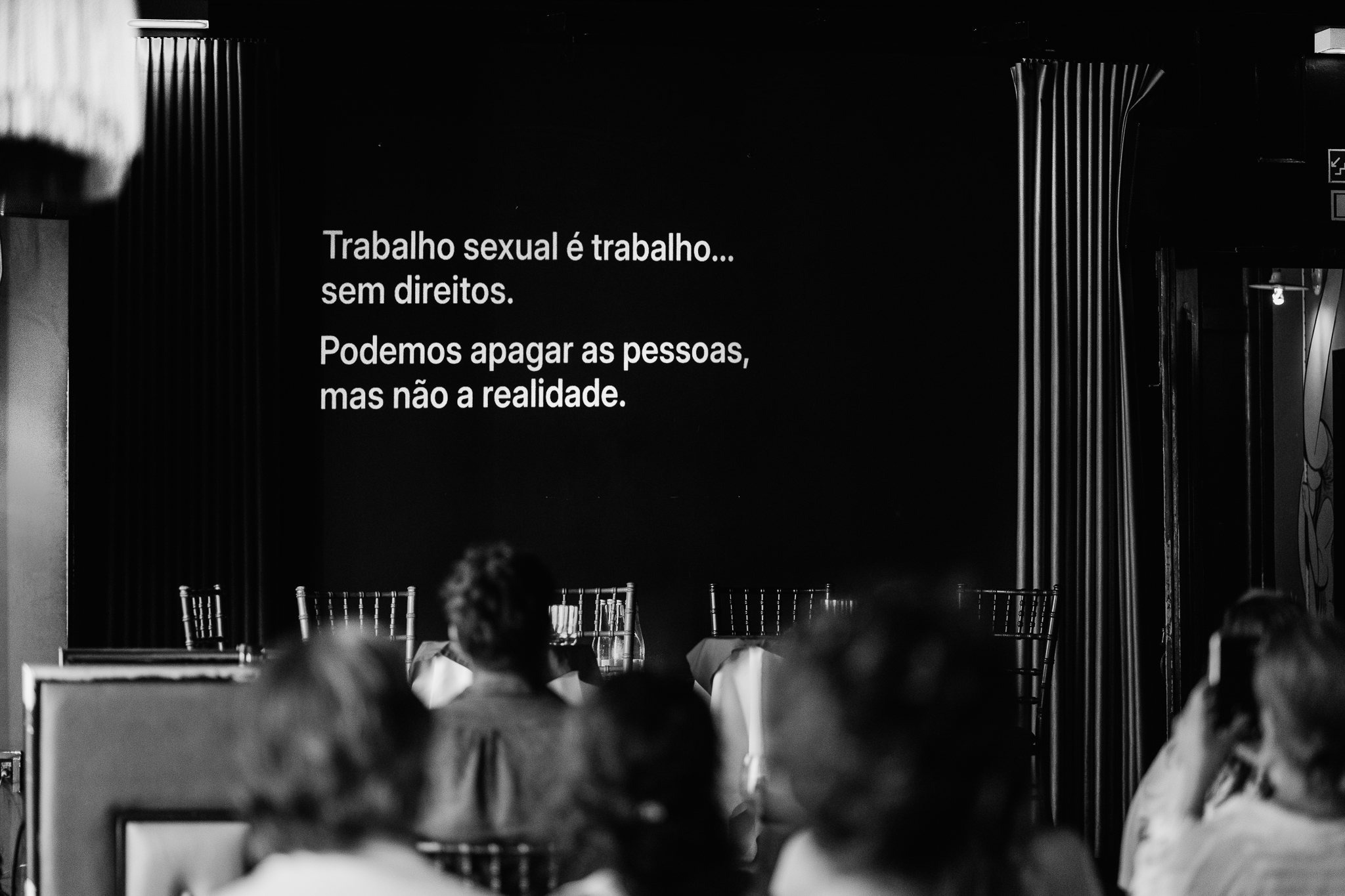Imagem a preto e branco com frases da campanha INVISÍVEIS projetadas numa tela de fundo escuro durante a mesa-redonda.