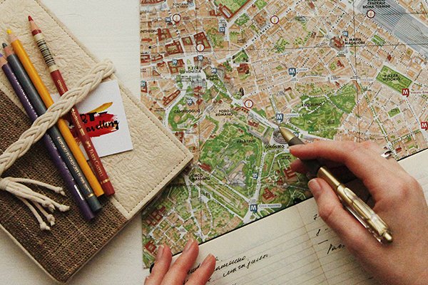 Pessoa a planear a sua viagem com um mapa, um caderno e alguns l&aacute;pis na mesa