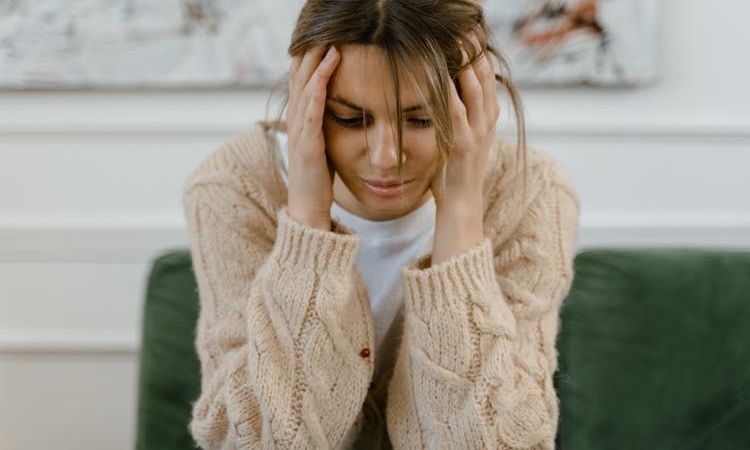 mulher sentada, debru&ccedil;ada sobre os joelhos, com as m&atilde;os a agarrar a cabe&ccedil;a e com cara de sofrimento