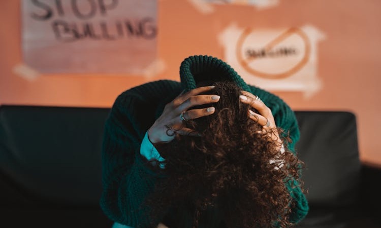 pessoa sentada, debru&ccedil;ada sobre os joelhos, com as m&atilde;os a segurar a nuca, com placares contra o bullying na parede atr&aacute;s dela