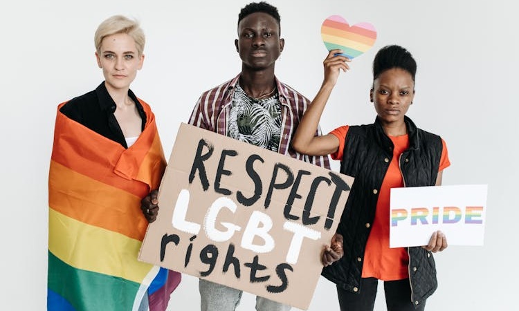 tr&ecirc;s pessoas a segurar cartazes em prol dos direitos LGBTQIAPN+, uma enrolada na bandeira da comunidade