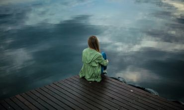 Mulher sentada num estrado de madeira a olhar para um lago, com um casaco verde vestido e a pensar nos sintomas de depressão.