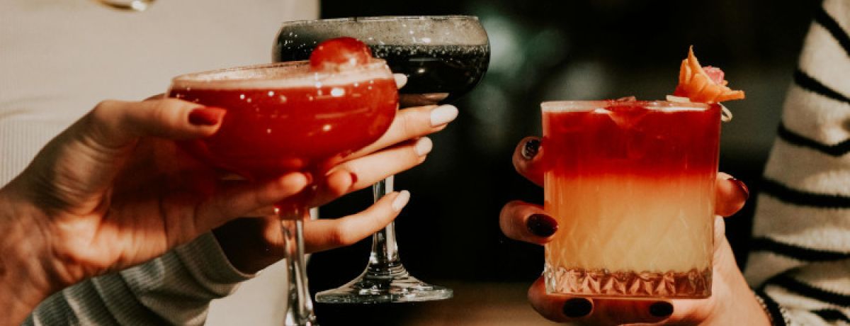 Mãos de 3 mulheres com unhas pintadas segurando bebidas para mostrar o significado de casa de alterne.