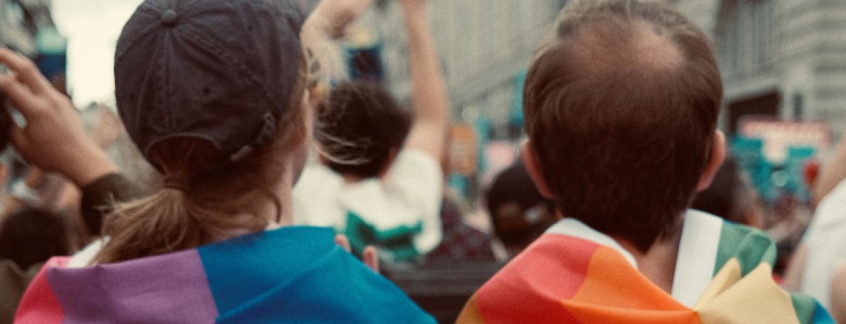 Pessoa assexual e outra pessoa pansexual de costas, com bandeiras multicoloridas, a olhar para multidão durante protesto.
