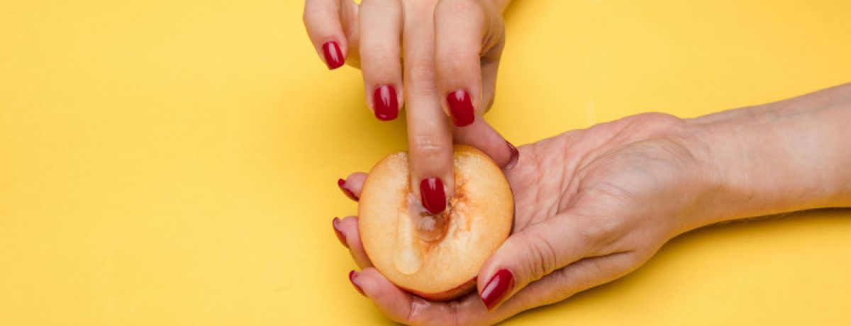 Mãos de mulher com unhas pintadas de vermelho a tocar pêssego com líquido viscoso a mostrar como observar o muco cervical no método do muco.