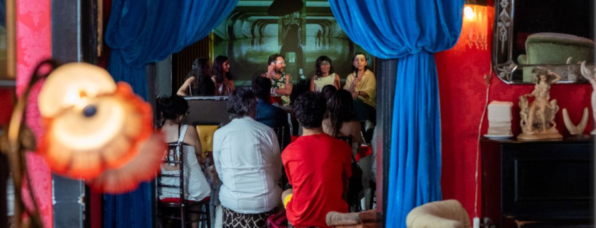 Pessoas de costas na Pensão Amor, com cortinas azuis a esconder parte da sala, durante mesa-redonda da Campanha Invisíveis.