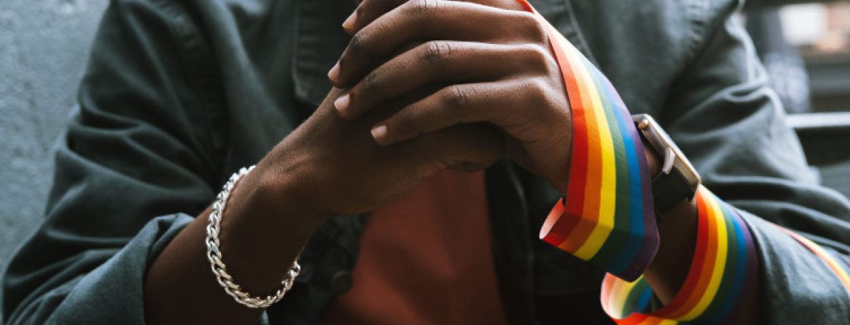 Close-up de mãos entrelaçadas com fita arco-íris a representar o orgulho LGBTQIAPN+ e o outing.