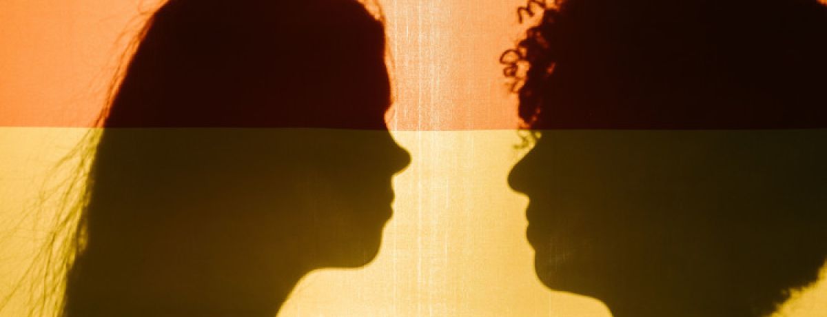 Perfis de duas pessoas a preto, um homem e uma mulher, com uma bandeira colorida como pano de fundo a representar a transição de género em Portugal.