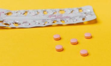 Tablete de uma pílula contraceptiva com cinco pílulas cor de rosa espalhadas num fundo amarelo.