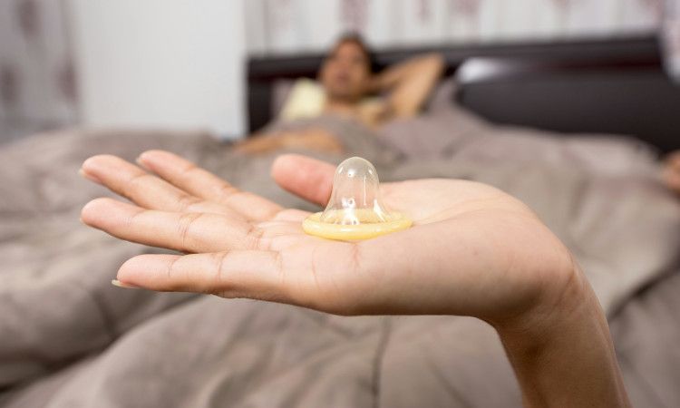 Mão de mulher a segurar camisinha com homem deitado na cama à espera e a pensar em como saber se o preservativo rompeu.