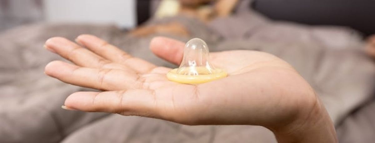 pessoa segura preservativo com a mão enquanto parceirx está na cama