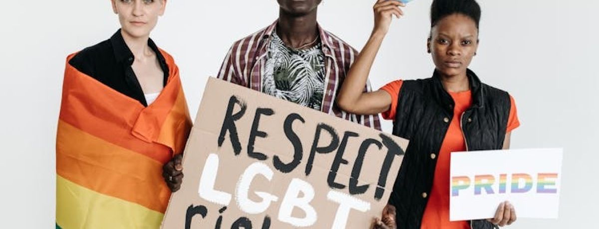 três pessoas a segurar cartazes em prol dos direitos LGBTQIAPN+, uma enrolada na bandeira da comunidade