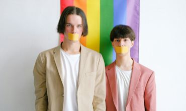 Dois jovens de camisolas brancas e fatos bege e rosa em frente a bandeira LGBTQIPN+ com fitas colas na boca a representar a homofobia.