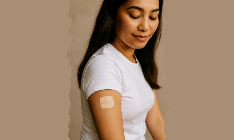 Mulher morena, com cabelos castanhos e um meio sorriso, a olhar para o adesivo contracetivo quadrado no braço.