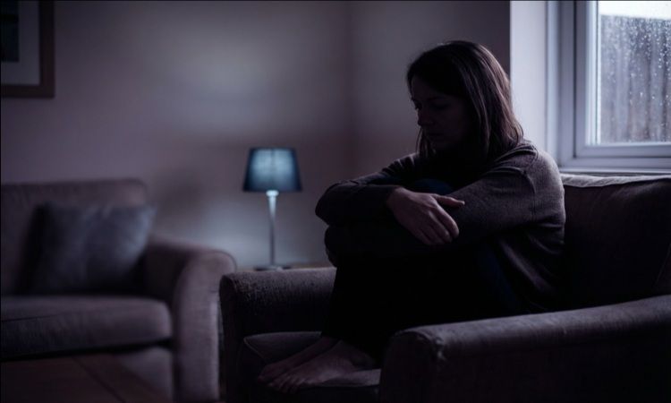 mulher sentada no sofá, enrolando os braços sobre os joelhos fletidos, em sala escura