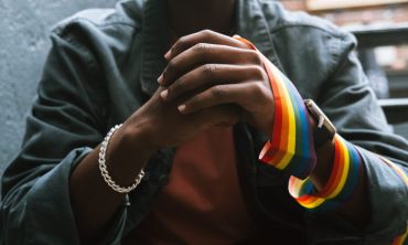 Close-up de mãos entrelaçadas com fita arco-íris a representar o orgulho LGBTQIAPN+ e o outing.