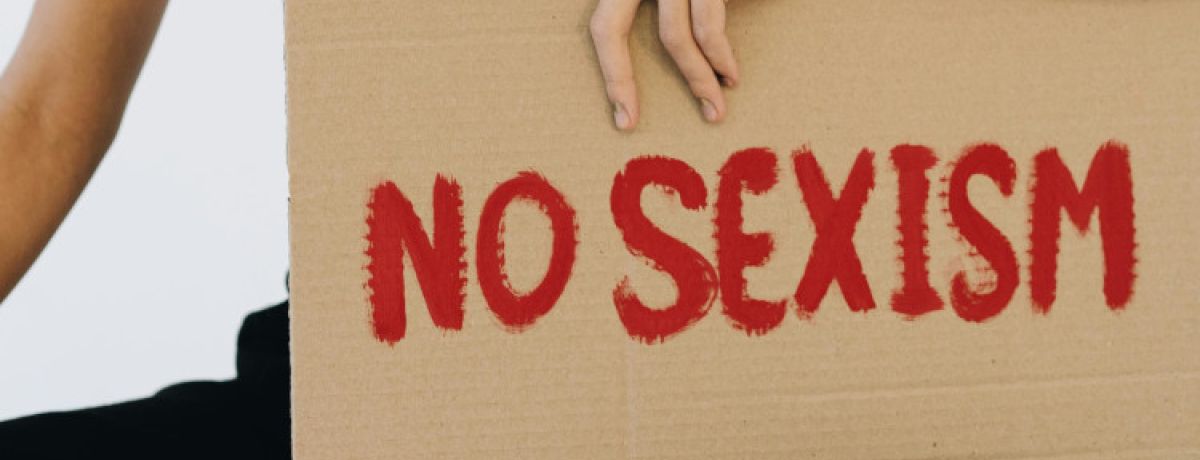 Mão de mulher a segurar cartaz de papel bege com as palavras "Não ao sexismo" em inglês escritas a vermelho.