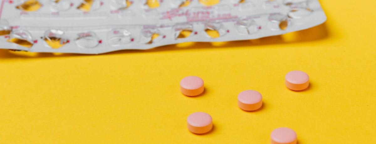 Tablete de uma pílula contraceptiva com cinco pílulas cor de rosa espalhadas num fundo amarelo.