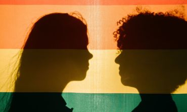 Perfis de duas pessoas a preto, um homem e uma mulher, com uma bandeira colorida como pano de fundo a representar a transição de género em Portugal.