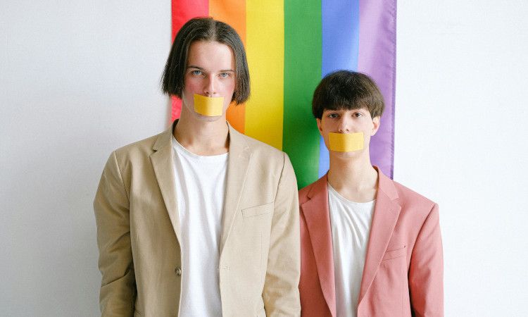 Dois jovens de camisolas brancas e fatos bege e rosa em frente a bandeira LGBTQIPN+ com fitas colas na boca a representar a homofobia.