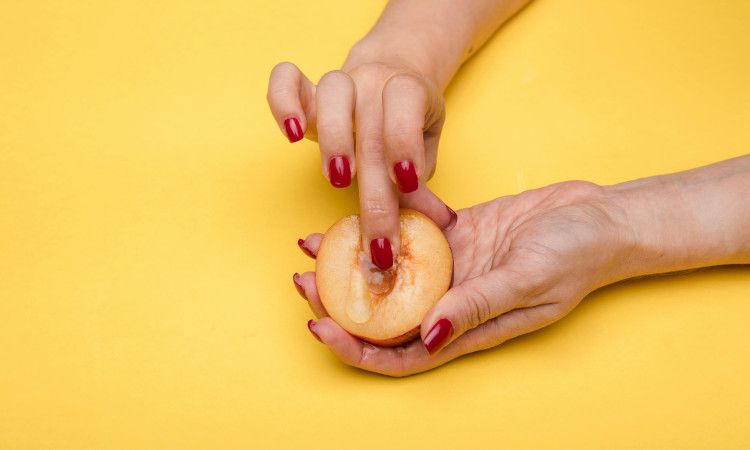 Mãos de mulher com unhas pintadas de vermelho a tocar pêssego com líquido viscoso a mostrar como observar o muco cervical no método do muco.
