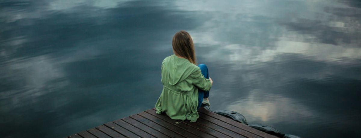 Mulher sentada num estrado de madeira a olhar para um lago, com um casaco verde vestido e a pensar nos sintomas de depressão.