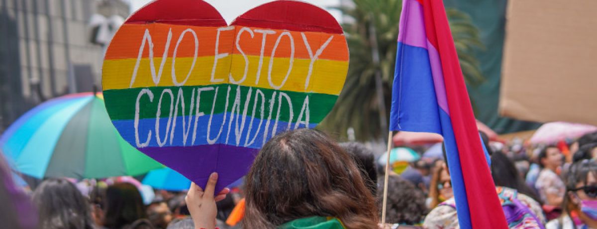 Pessoas na rua com uma mulher em destaque, a segurar um cartaz colorido em forma de coração que diz "Não estou confusa" em espanhol, para ilustrar o que é ser bissexual.