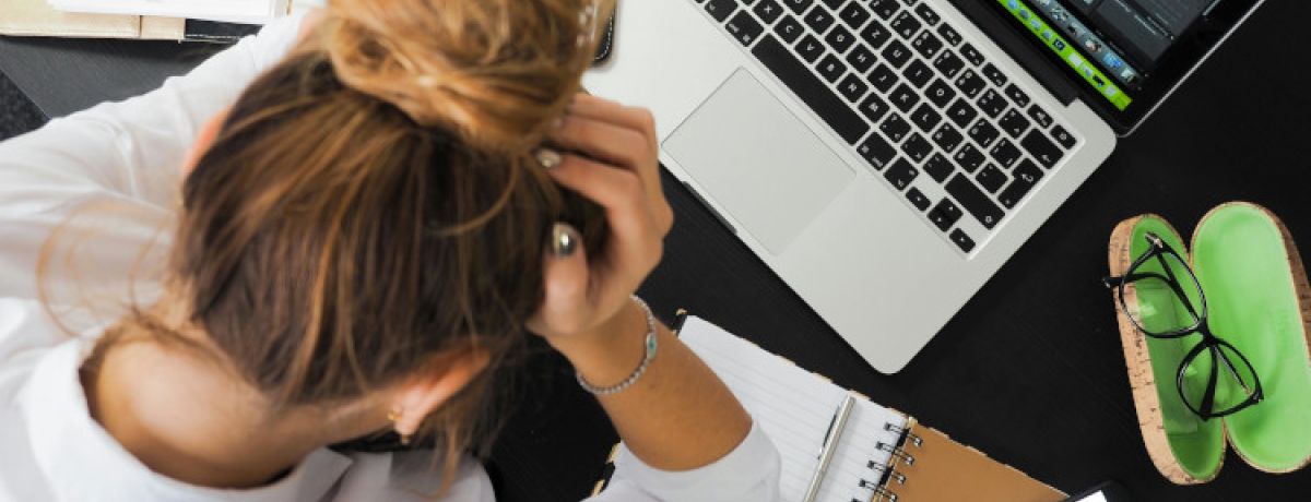 Mulher com o cabelo apanhado e as mãos a segurar a cabeça em frente ao computador com sintomas de ansiedade.
