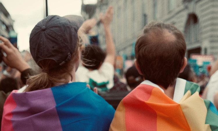 Pessoa assexual e outra pessoa pansexual de costas, com bandeiras multicoloridas, a olhar para multidão durante protesto.