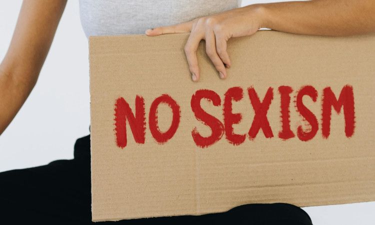Mão de mulher a segurar cartaz de papel bege com as palavras "Não ao sexismo" em inglês escritas a vermelho.