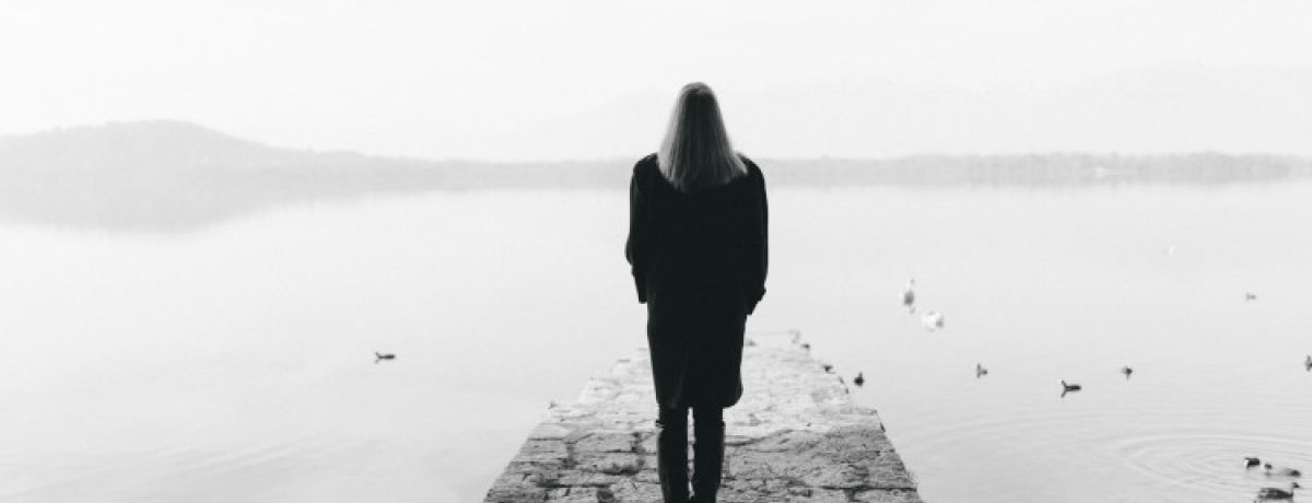 Fotografia a preto e branco com mulher de corpo inteiro, cabelos compridos, a olhar para um lago a partir de um passadiço e a pensar no estigma e no preconceito social.