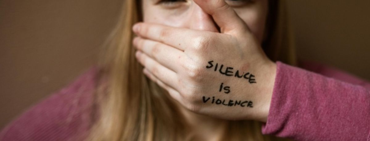 jovem vítima de violência no namoro tapa a boca com a mão na qual tem escrita a mensagem silêncio é violência