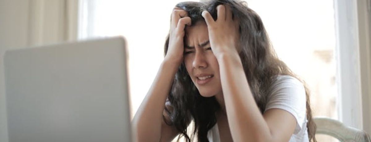 mulher sentada na secretária, à frente do computador, com as mãos na cabeça e cara triste, revoltada