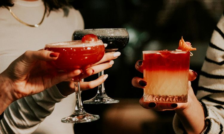 Mãos de 3 mulheres com unhas pintadas segurando bebidas para mostrar o significado de casa de alterne.