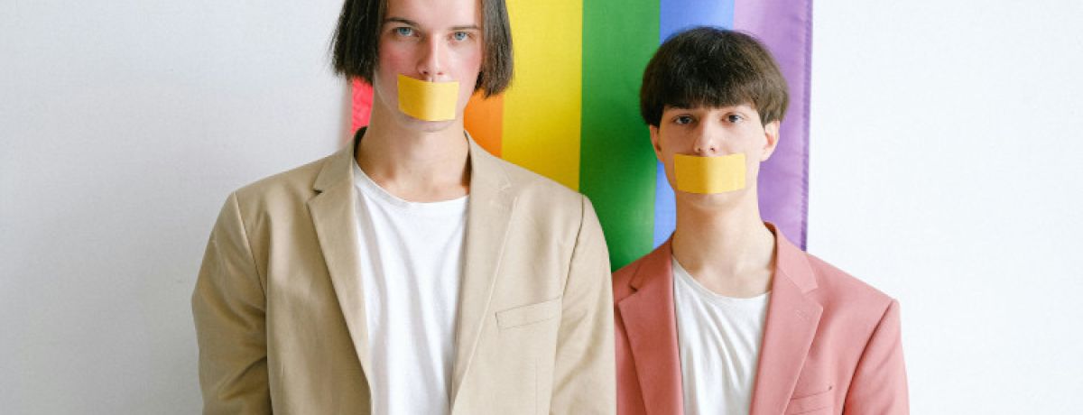 Dois jovens de camisolas brancas e fatos bege e rosa em frente a bandeira LGBTQIPN+ com fitas colas na boca a representar a homofobia.