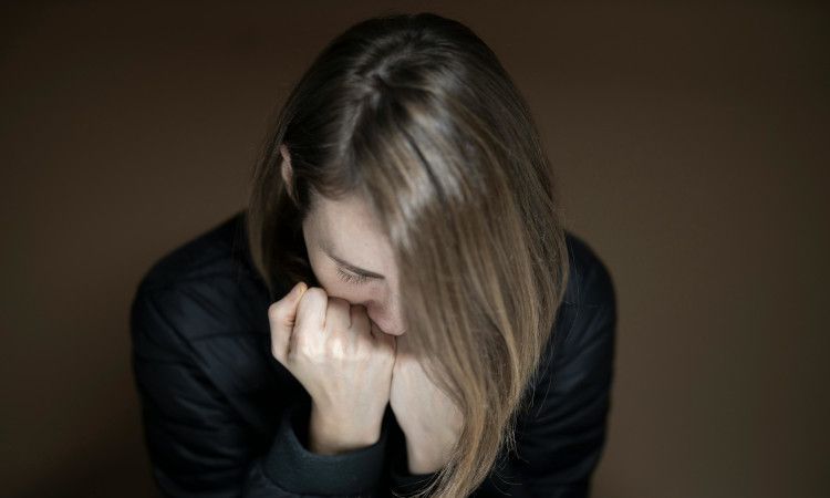 Mulher de camisola preta e cabelo comprido castanho com as mãos junto à boca e a cabeça debruçada durante ataque de ansiedade.