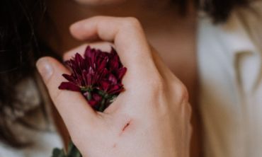Mão de mulher a segurar flor num vermelho escuro com um arranhão em evidência a sinalizar um dos vários tipos de violência que existem.