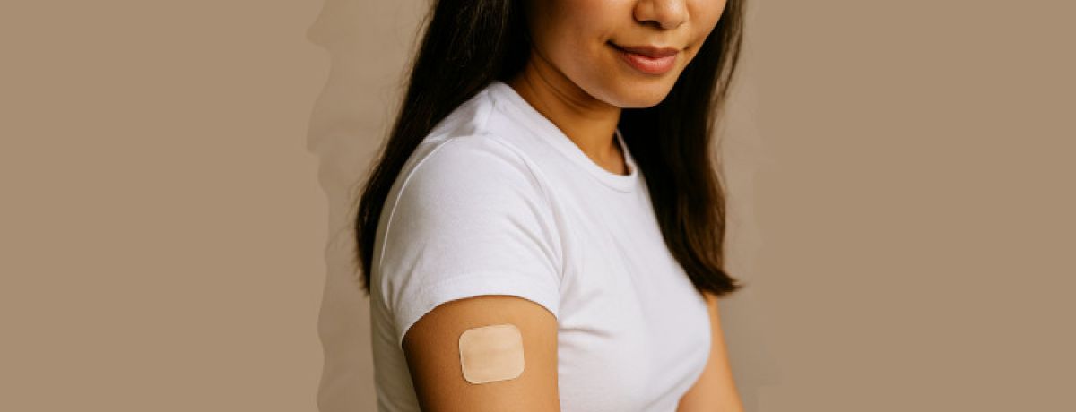 Mulher morena, com cabelos castanhos e um meio sorriso, a olhar para o adesivo contracetivo quadrado no braço.