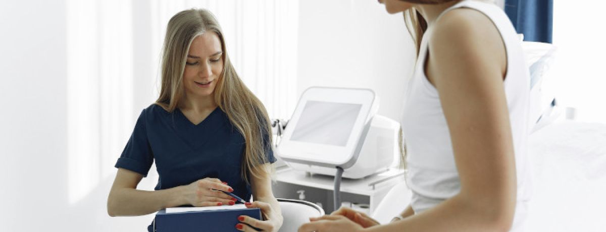 Profissional de saúde de unhas pintadas de vermelho e camisola e calças azuis escuras a falar com mulher vestida de branco sobre o HPV.