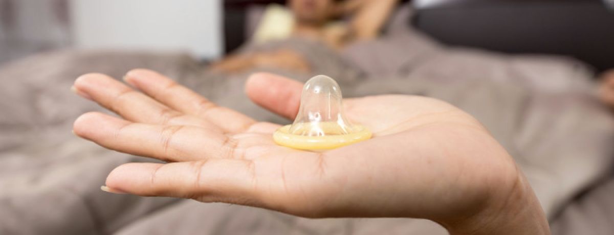 Mão de mulher a segurar camisinha com homem deitado na cama à espera e a pensar em como saber se o preservativo rompeu.