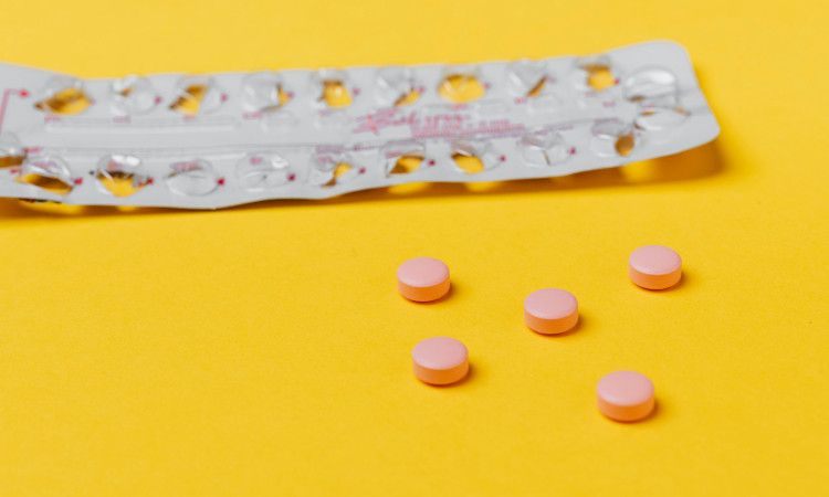 Tablete de uma pílula contraceptiva com cinco pílulas cor de rosa espalhadas num fundo amarelo.