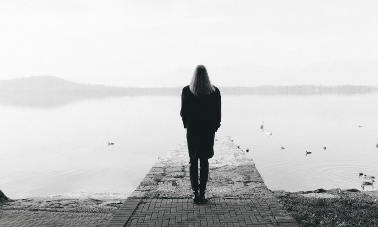 Fotografia a preto e branco com mulher de corpo inteiro, cabelos compridos, a olhar para um lago a partir de um passadiço e a pensar no estigma e no preconceito social.