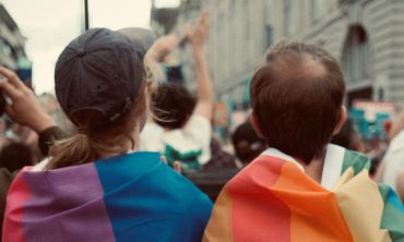 Pessoa assexual e outra pessoa pansexual de costas, com bandeiras multicoloridas, a olhar para multidão durante protesto.