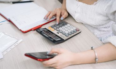 Tronco de mulher com blusa branca e unhas pintadas de azul claro a mexer no telemóvel e numa calculadora enquanto trabalhador independente Segurança Social.