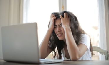 mulher sentada na secretária, à frente do computador, com as mãos na cabeça e cara triste, revoltada