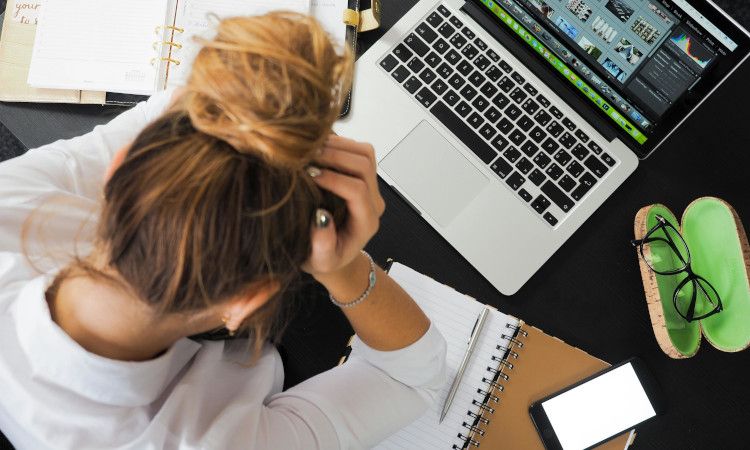 Mulher com o cabelo apanhado e as mãos a segurar a cabeça em frente ao computador com sintomas de ansiedade.